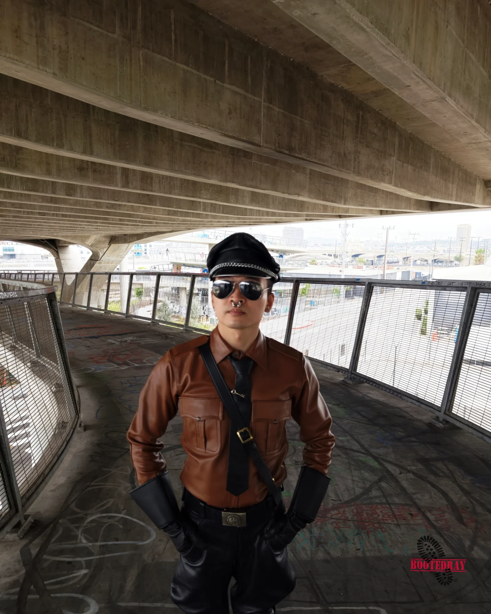 Man in brown leather uniform and black boots standing under a concrete bridge.