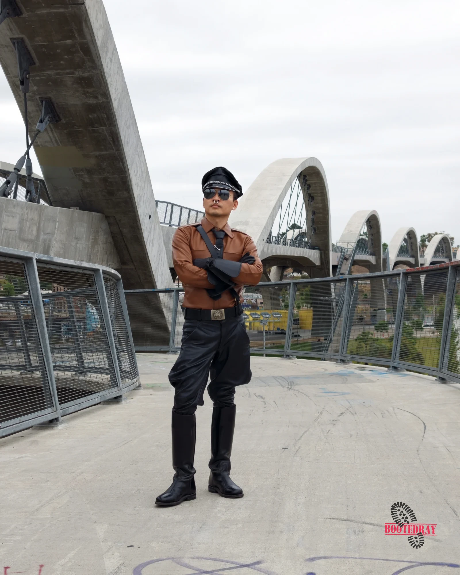 Man in brown leather uniform and black boots standing with arms crossed.