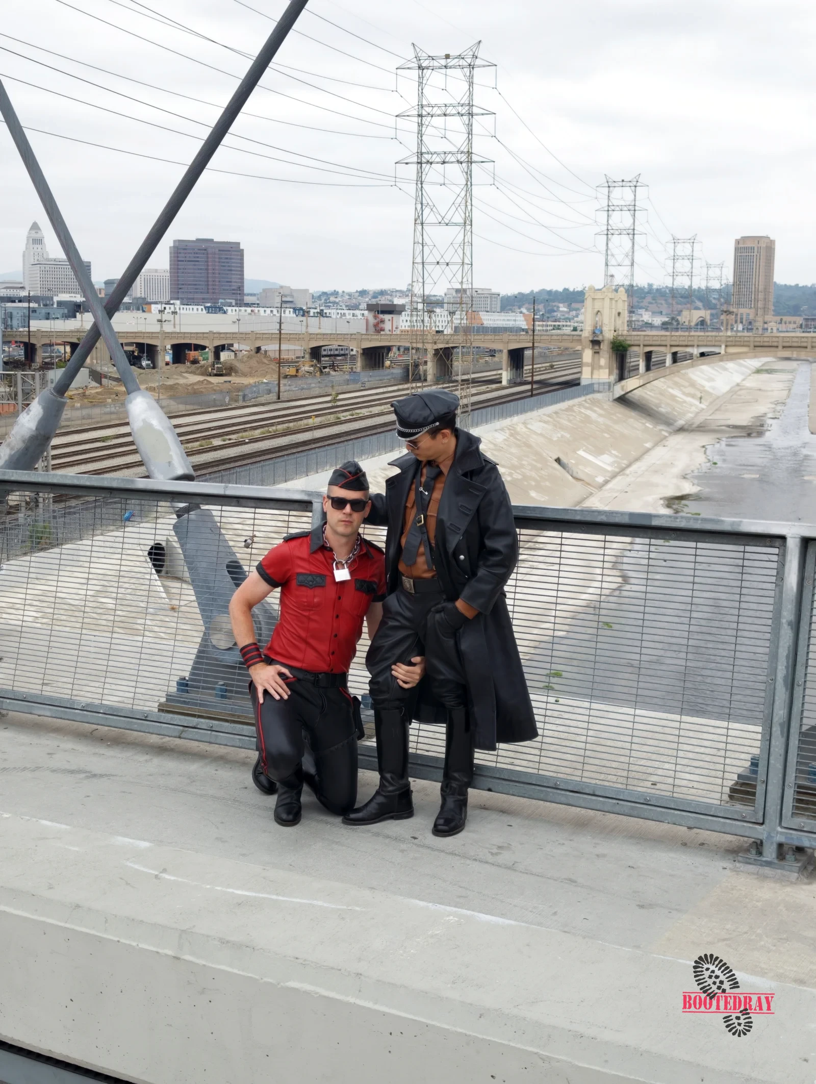 Two men in leather and red shirts posing on a bridge.