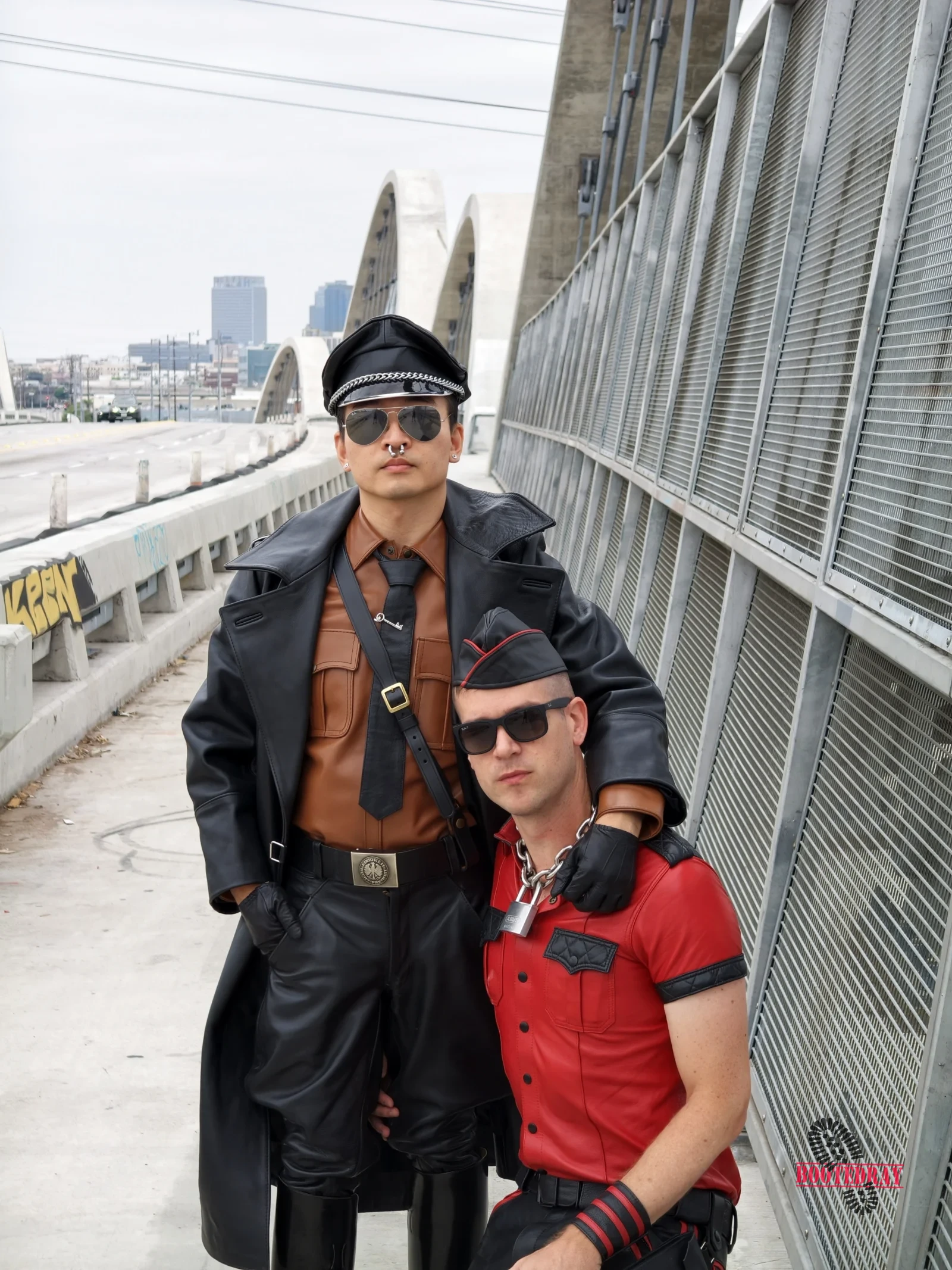 Man in black leather uniform and man in red shirt posing on a bridge.