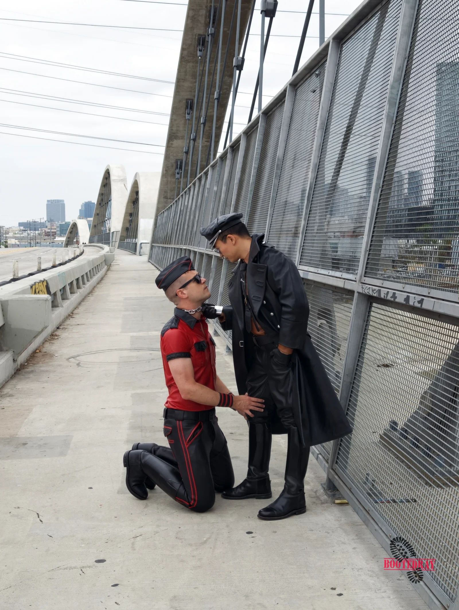 Two men in leather gear on a bridge walkway. One kneels in red shirt, one stands in black coat holding a chain.