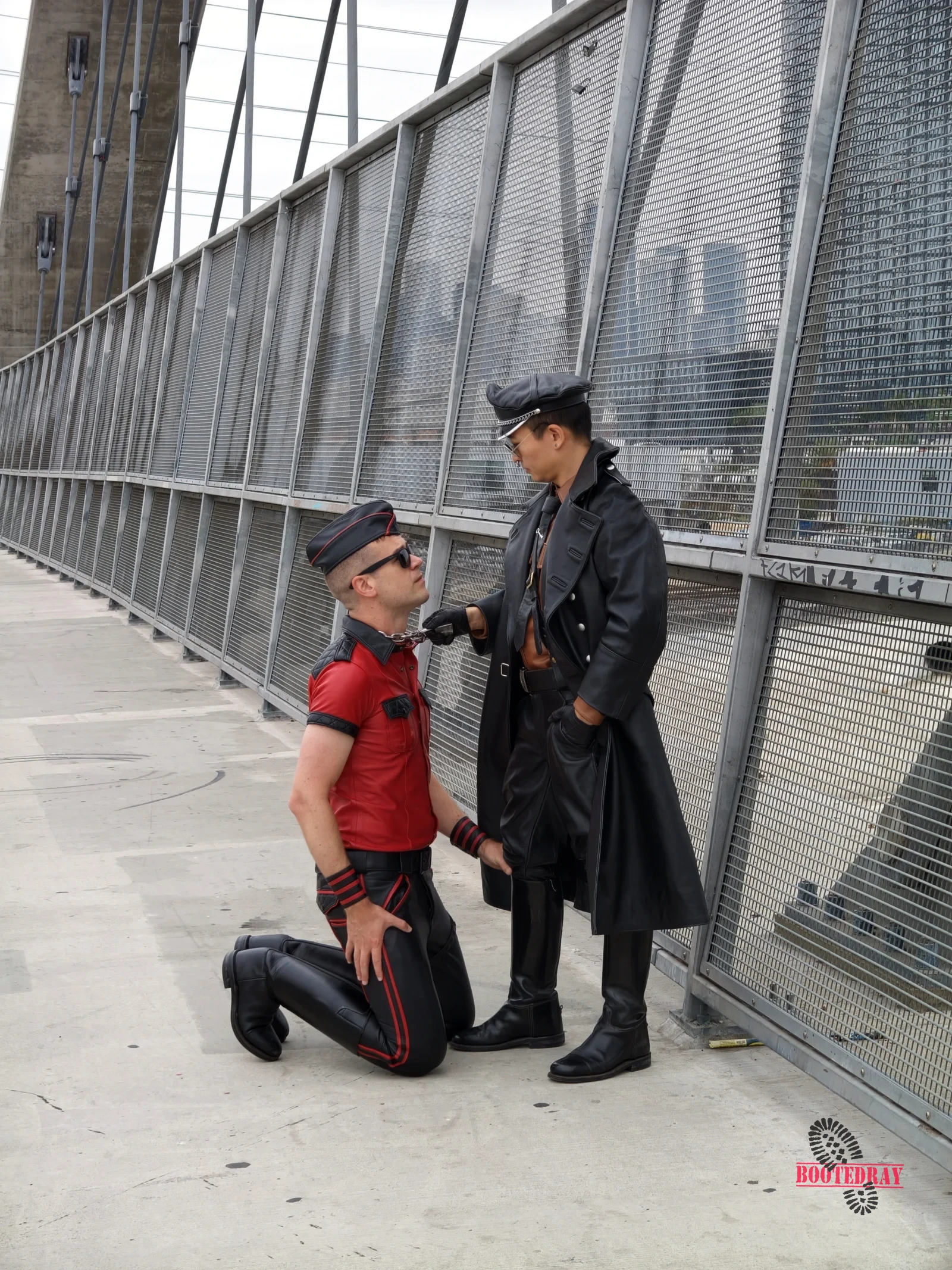 Close up of two men in leather gear on a bridge. One kneels in red, one stands in black holding a chain.