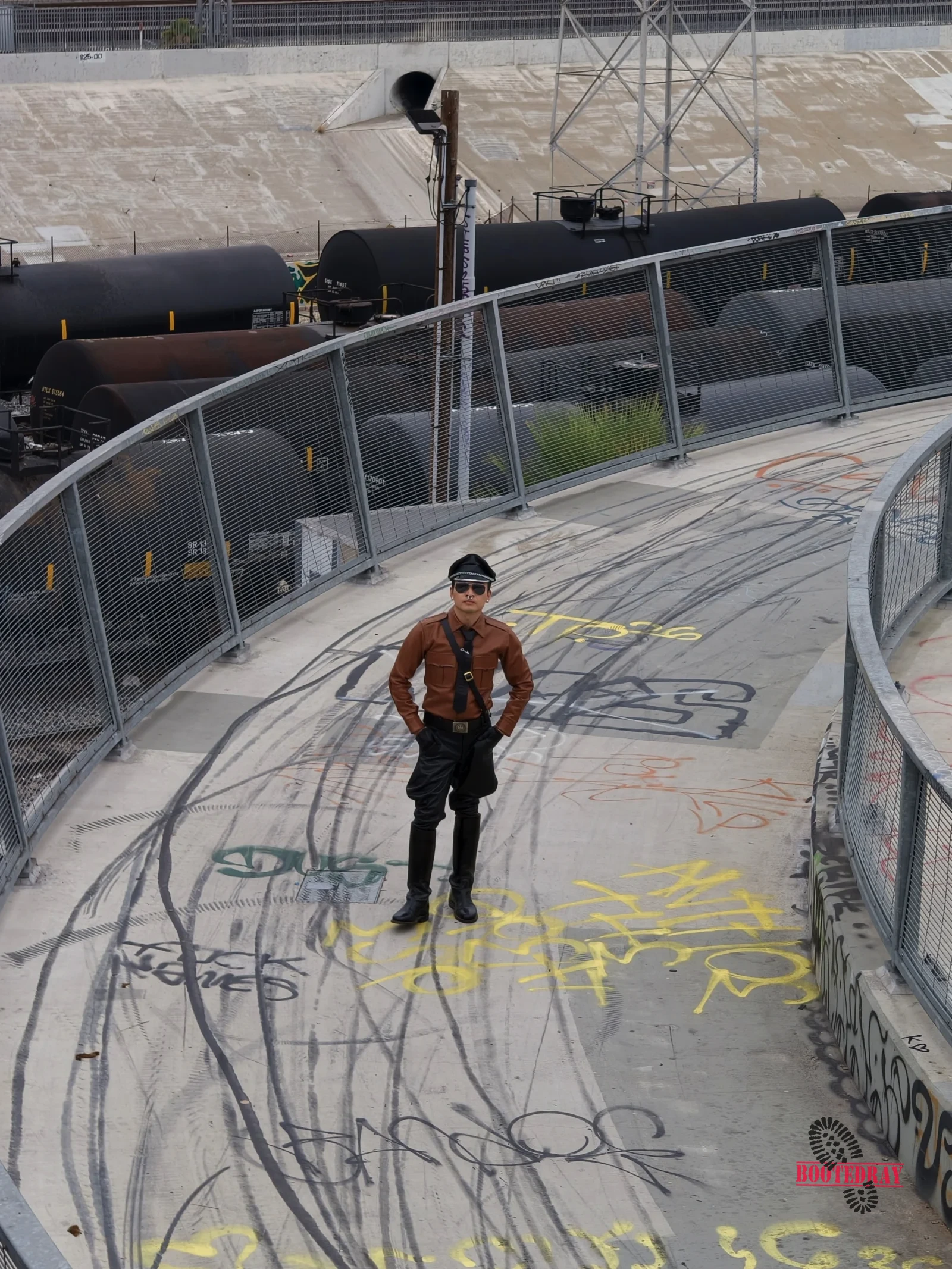 A man in a brown leather shirt stands on a graffiti-covered bridge walkway.